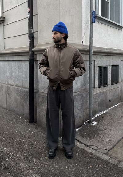 Hombre con gorro azul, chaqueta marrón con cremallera, pantalones anchos grises y botas negras, de pie en la acera de una ciudad cerca de la esquina de un edificio.