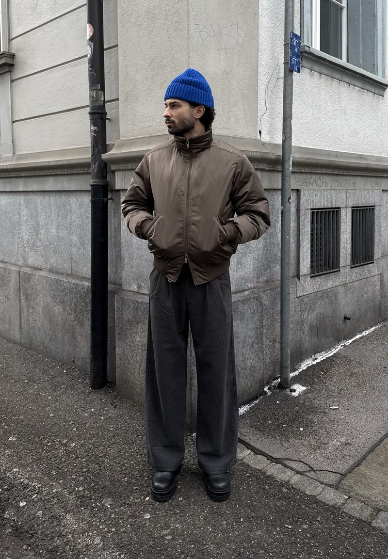 Homme portant un bonnet bleu, une veste marron à fermeture éclair, un pantalon large gris et des bottes noires, debout sur un trottoir de la ville près d'un coin de bâtiment.