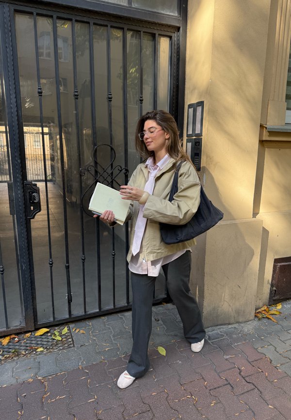A person wearing a light beige jacket, a pink shirt, and dark trousers is carrying a green notepad. They have clear glasses and are holding a black bag. The background includes a gate.