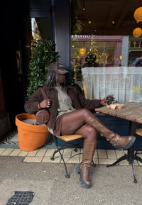 Γυναίκα με πλεξούδες, φορώντας καφέ μπότες, μπουφάν, φούστα και καπέλο, κάθεται χαλαρή έξω από καφετέρια κρατώντας ένα ποτήρι καφέ για να το πάρει μαζί της.