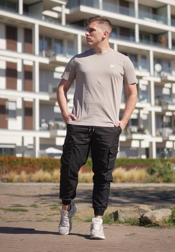 Joven con camiseta beige y pantalones cargo negros caminando al aire libre en una zona urbana con edificios de apartamentos al fondo.