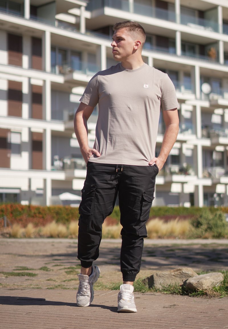 Jeune homme en t-shirt beige et pantalon cargo noir marchant en extérieur dans un quartier urbain avec des immeubles en arrière-plan.
