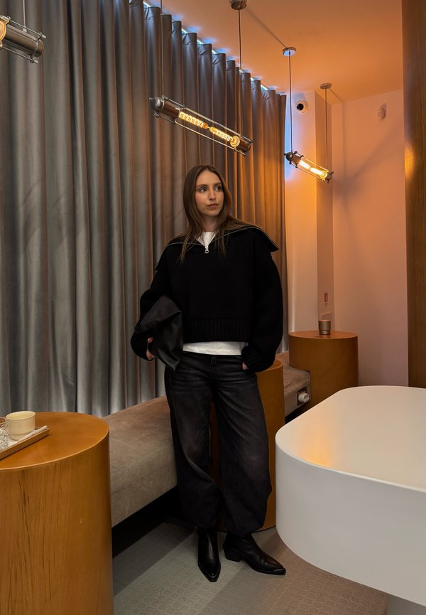 Kvinne i svart genser og bukser som holder en clutch, stående i et moderne rom med tremøbler, grå gardiner og varme hengende lys.