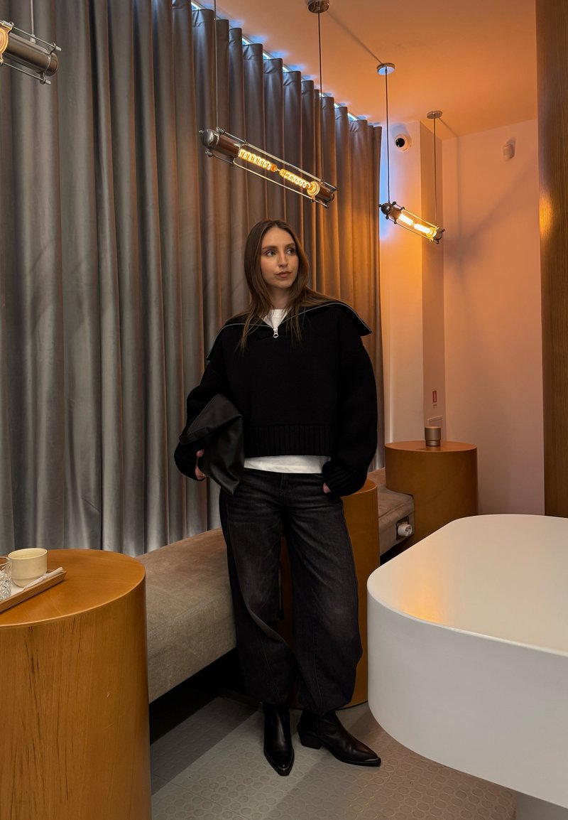 Žena v čiernom svetri a nohaviciach drží kabelku, stojí v modernej miestnosti s dreveným nábytkom, sivými závesmi a teplým závesným osvetlením.