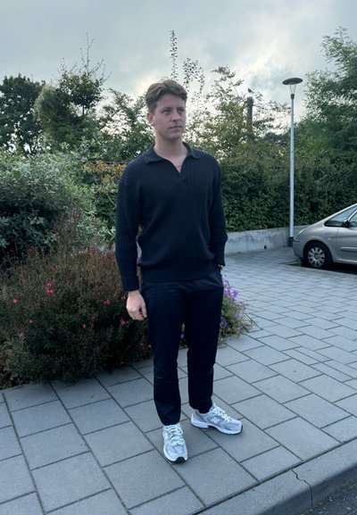 Suéter de polo de manga larga de canalé oscuro, pantalones negros a medida y zapatillas gris. De pie sobre una superficie pavimentada cerca de vegetación y flores.