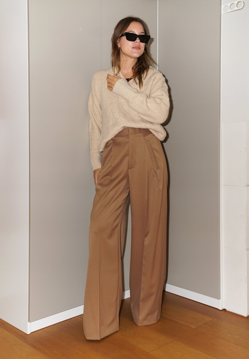 Beige neulepusero, joka on yhdistetty korkeavyötäröisiin, leveisiin ruskeisiin housuihin. Mallilla on mustat aurinkolasit neutraalin taustan edessä.