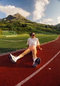 Sportinė avalynė su juodais padais, balta tinklelio viršūne. Nešiojantis asmuo vilki baltą marškinėlį, rožinius šortus, sėdi ant raudono bėgimo tako su žolės fono.