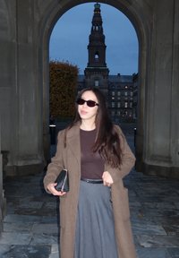 Brauner Überwurf, passendes braunes Oberteil, grauer Rock, schwarze Sonnenbrille, hält eine schwarze Clutch, vor einem historischen Gebäude und einem Bogen.