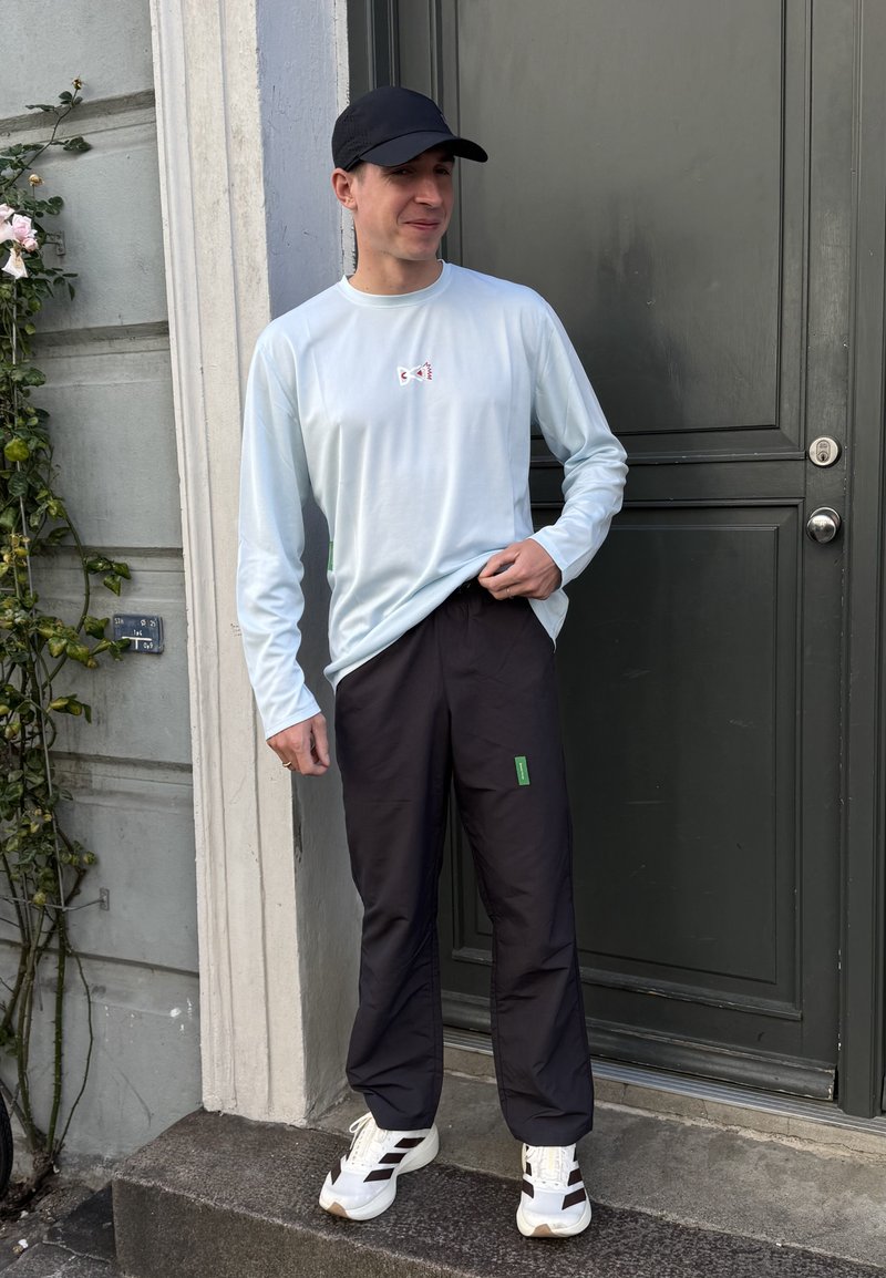 Un hombre con una gorra negra, una camisa azul claro de manga larga, pantalones oscuros y zapatillas blancas está junto a una puerta, levantándose ligeramente la camisa.