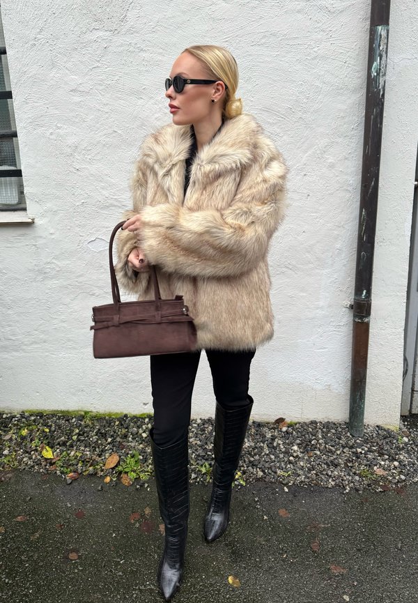 Pelskåpe i lys beige, oversized passform. Svarte leggings, knelange spisse støvler med mønstret tekstur. Brun håndveske med strukturert design.