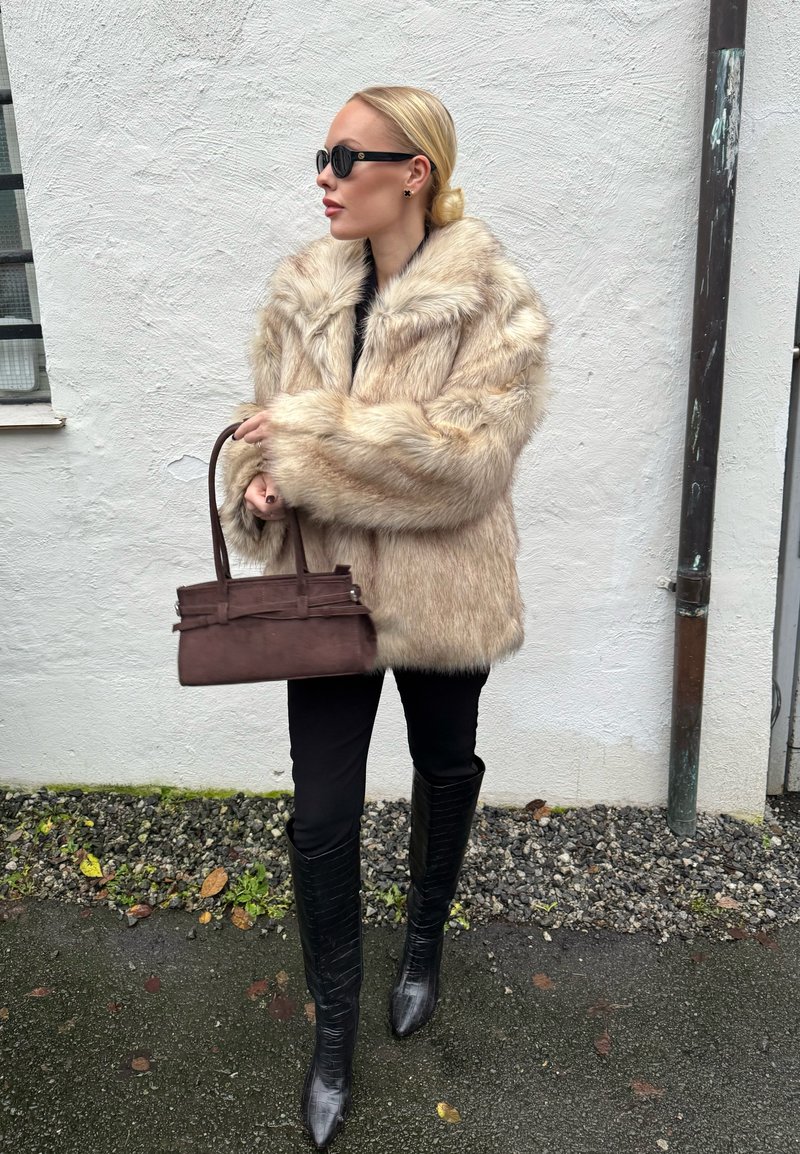 Plašč iz umetnega krzna v svetlo bež barvi, ohlapna oblika. Črne legice, škornji do kolen z nabrušenim koncem in teksturiranim vzorcem. Rjava torbica s strukturiranim dizajnom.