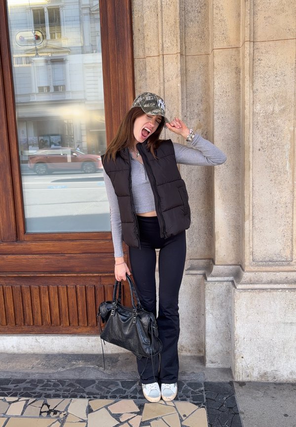 Une femme portant une casquette camouflage, un gilet noir et un pantalon noir pose devant un bâtiment en pierre, tenant un sac à main noir et soulevant sa casquette.