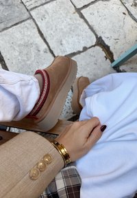 Personne portant des chaussures en suede beige avec des bordures rouges et blanches, un pantalon blanc, une veste beige avec des boutons, des bracelets en or et du vernis à ongles noir, assise sur un sol en pierre.