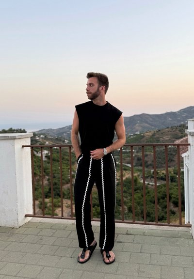 Top negro sin mangas combinado con pantalones negros que presentan rayas laterales onduladas en blanco. El calzado son chanclas negras; de pie contra un fondo escénico.