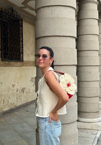 Λευκή υφασμάτινη μπλούζα με κορδόνια στα πλάγια, ανοιχτό γαλάζιο τζιν παντελόνι και μια τσάντα με κόκκινη πλάτη από υφαντό υλικό με λεπτομέρεια από κρεμ λουλούδι.