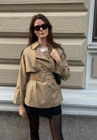 Beigefärgad dubbelknäppt trench coat med bälte, axelremmar och vida ärmar. Bärs över en svart kjol, matchad med svarta solglasögon och en liten väska.