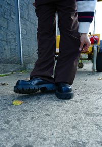 Fekete platform cipők tömör talppal, sima felülettel és lekerekített orral, sötétbarna nadrággal viselve, egy beton kültéri környezetben.