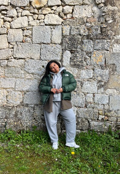Chaqueta acolchada verde sobre una sudadera gris, pantalones de chándal grises y zapatillas blancas. Posando frente a una pared de piedra texturizada con hierba verde abajo.