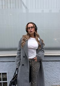 Szürke gyapjú kabát nagy gombokkal, fehér szűk szabású ing, és leopárdmintás szűk farmernadrág. Fekete fejhallgató, hosszú hullámos haj, minimalista fekete táska.