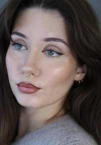Close-up of a face with light brown skin, blue eyes, winged eyeliner, a gold septum piercing, and glossy bronze lips. Brown hair and soft texture.
