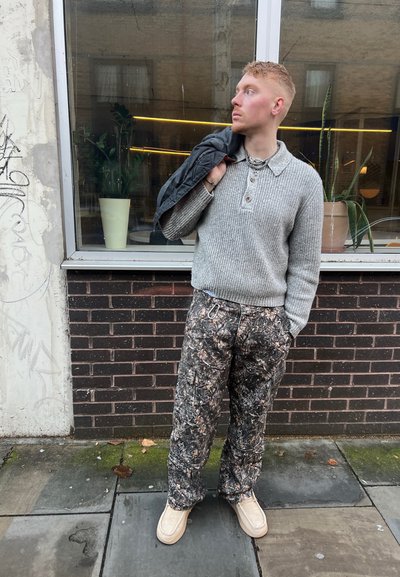 Suéter gris de canalé con un cuello tipo polo, pantalones de camuflaje y zapatos tipo slip-on de color crema, de pie cerca de una ventana con plantas.