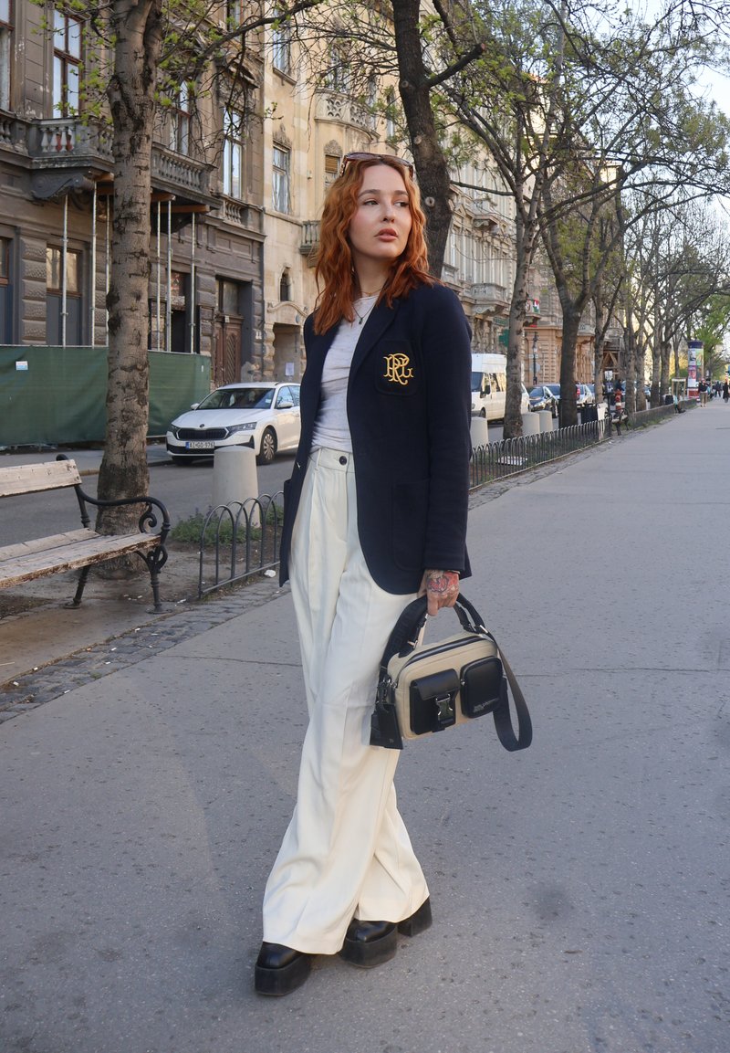 Junge Frau mit roten Haaren, die einen dunkelblauen Blazer und weiße Hosen trägt, eine schwarz-beige Handtasche bei sich hat und auf einem Bürgersteig in der Stadt geht.