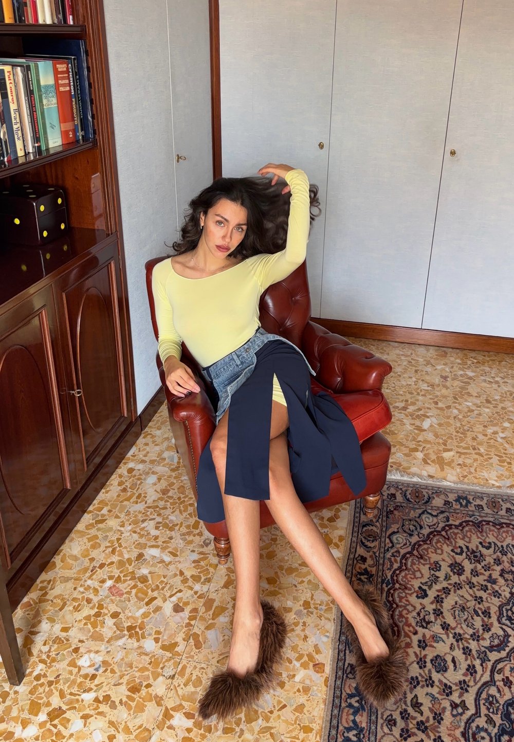 Žlutý dlouhý rukáv, džínsová sukně s námořnickými akcenty a huňaté hnědé mules. Sedí na červené kožené židli na pozadí vzorovaného koberce.