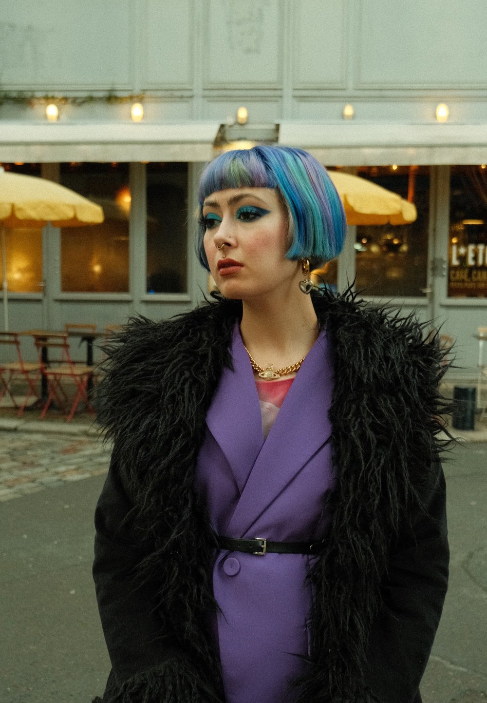 Manteau violet avec col en fausse fourrure noire, coiffure bob colorée, fard à paupières bleu audacieux, collier en or superposé. Cadre de café en plein air.