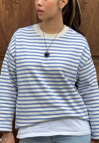 Chemise blanche à manches longues avec des rayures horizontales bleues, coupe décontractée, portée par-dessus un t-shirt blanc. Un collier avec un pendentif en forme de cœur est porté.