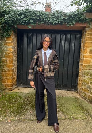 Joven con cárdigan marrón con estampado, camisa blanca, corbata marrón, pantalones negros de pierna ancha y gafas, posando frente a una puerta de garaje negra.