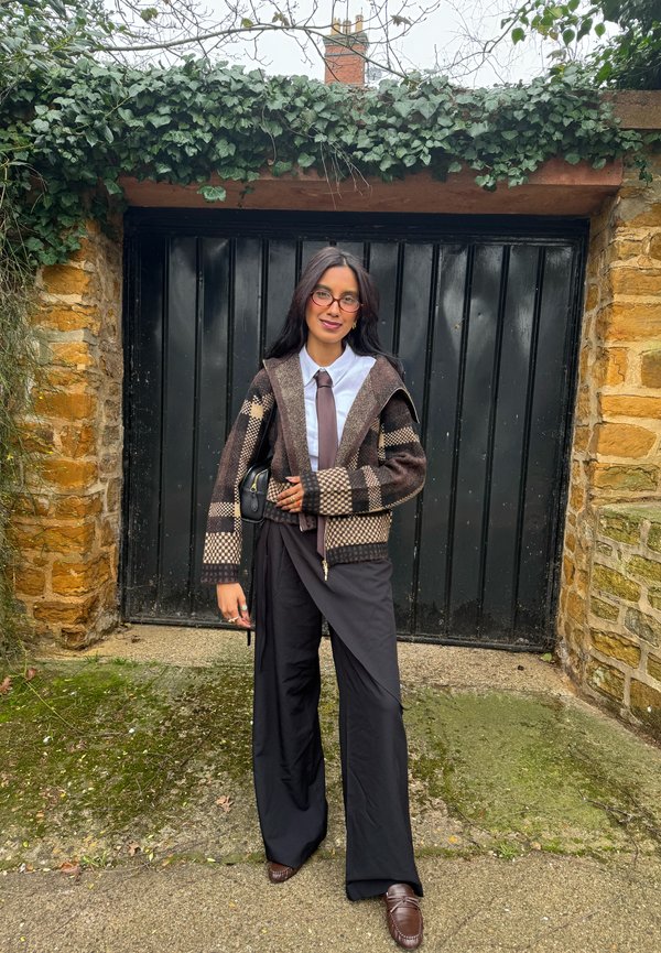 Ung kvinne iført brun mønstret cardigan, hvit skjorte, brun slips, svarte bukser med vide ben og briller står foran en svart garasjeport.