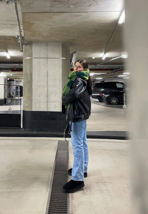 Černá kožená bunda, zeleno-modrý šátek, světle modré džíny, černé kotníkové boty. Stojí v parkovacím garáži s betonovými sloupy.