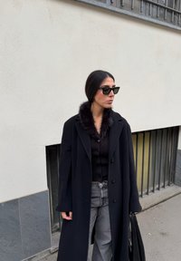 Manteau en laine noire avec col en fourrure, porté sur un pull noir à côtes, associé à un pantalon large gris et des lunettes de soleil noires. Sac à main noir inclus.