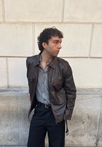 Mężczyzna z kręconymi włosami, ubrany w ciemnobrązową skórzaną kurtkę, pasiastą koszulę na guziki i czarne spodnie, stojący na tle beżowej kamiennej ściany.