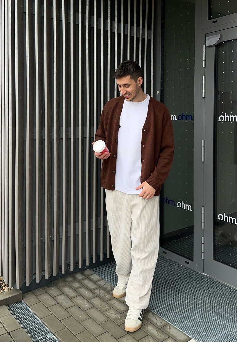 Cardigan marron sur un t-shirt blanc, pantalon en velours côtelé beige, tenant une tasse de café blanche, debout près d'une entrée moderne avec des lames verticales.