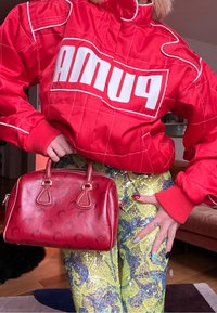Personne portant une veste rouge Puma et un pantalon jaune à paillettes, tenant un sac à main en cuir rouge avec des motifs de croissant de lune.