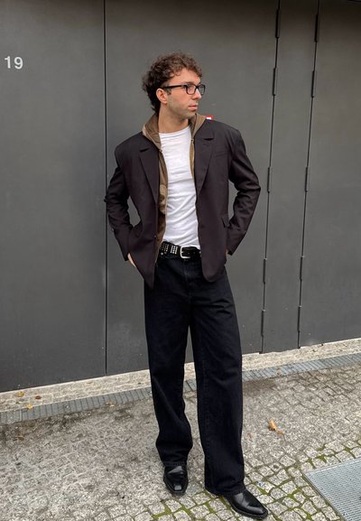 Blazer negro sobre una sudadera marrón, camiseta blanca y jeans de pierna ancha negros. Cinturón negro con tachuelas plateadas, zapatos negros. Atuendo urbano casual.