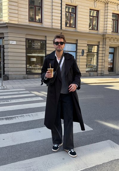 Hombre con gabardina negra y gafas de sol sostiene una taza de café mientras cruza una calle de la ciudad cerca de un edificio de piedra con grandes ventanas.