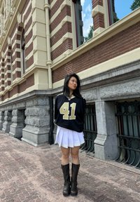 Nero maglietta NFL con numero 41 dorato, abbinata a una gonna bianca plissettata con rifiniture gialle e stivali al ginocchio marroni con fibbie.