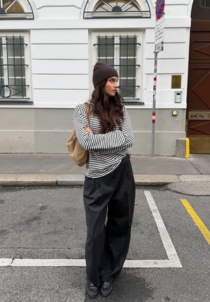 Camisa de manga larga a rayas blancas y negras, pantalones anchos de rayas negras, gorro de punto marrón y bolso beige al hombro. Entorno urbano callejero.