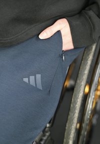 Main reposant dans la poche d'un pantalon Adidas bleu marine à côté de la roue d'un fauteuil roulant, portant un haut à manches longues noir.