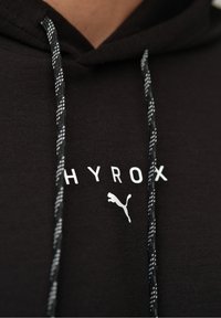 Czarna bluza z kapturem z kieszenią kangurzą, z białym logo "HYROX" i grafiką puma. Grube sznurki z wzorem w czarno-białe paski.