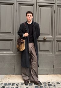 Melna garā mēteli virs tumša džempera, bēšas bikses un brūnas kurpes; tur rokā brūnu un bēšu somu; stāv pie teksturētas pelēkas durvis.