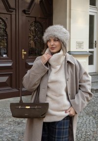 Manteau en laine brun clair porté sur un pull à col roulé crème, associé à un sac à main en daim marron avec un accent doré, devant des portes en bois ornées.