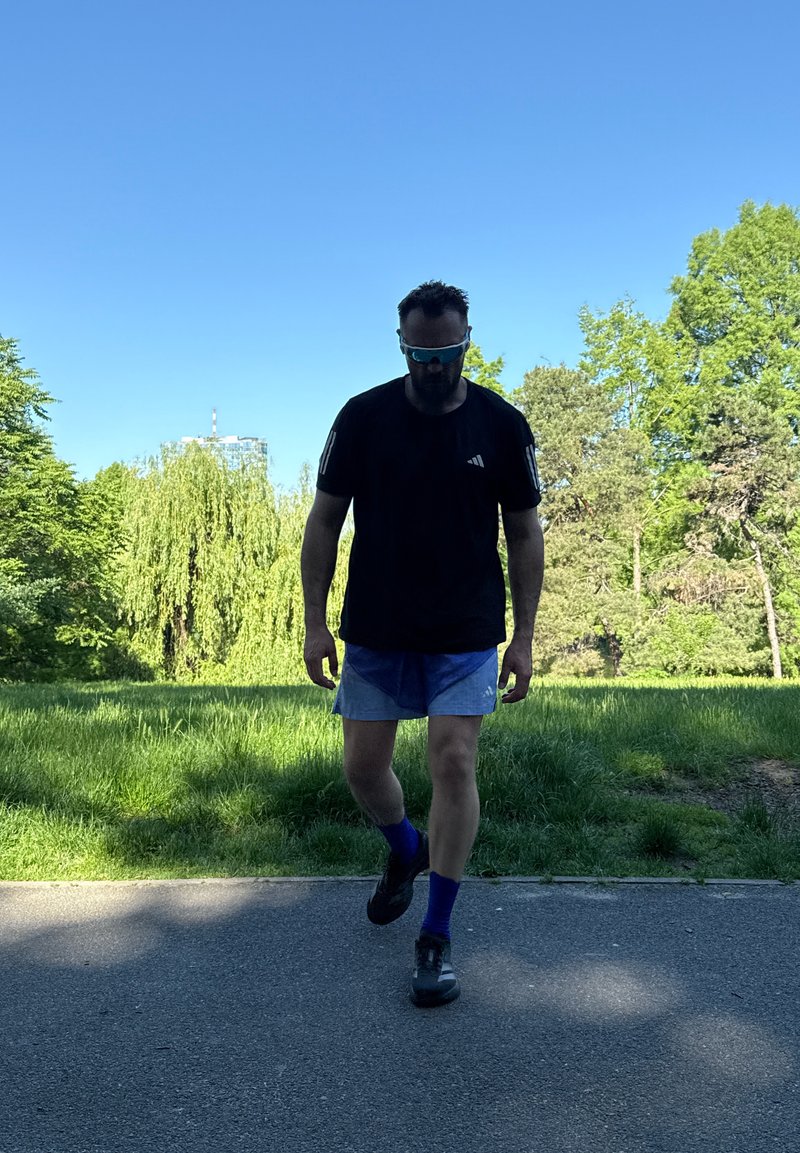 Homme portant des lunettes de soleil, une chemise noire, un short bleu et des chaussettes bleues marchant sur un chemin avec des arbres verts et un ciel bleu clair en arrière-plan.