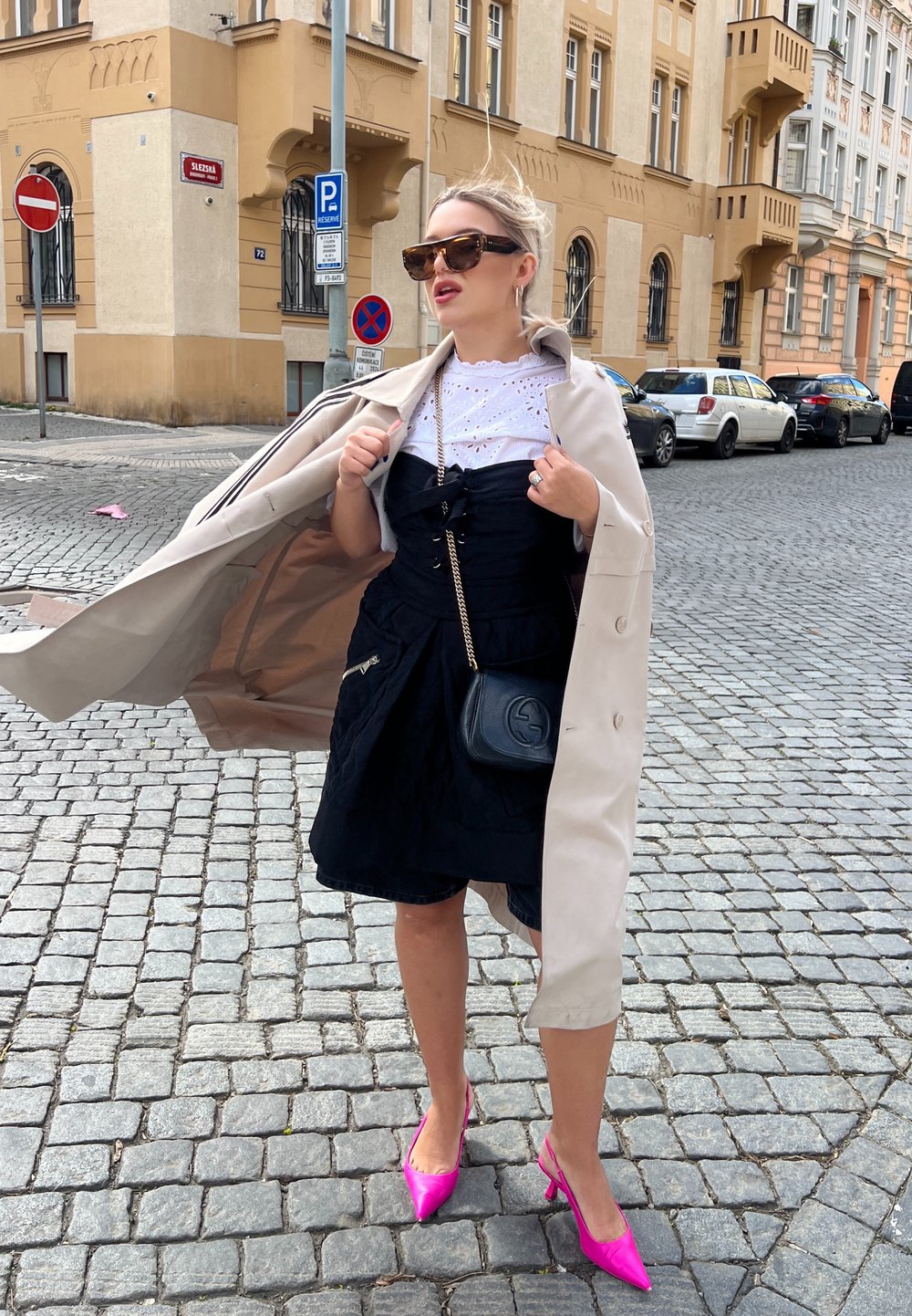 Vestido negro con fruncidos, blusa blanca de ojales, abrigo beige, tacones rosados de punta, y un bolso cruzado negro. Fondo de calle de adoquines.