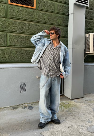 Chaqueta de denim sobre una camisa a cuadros gris, combinada con pantalones vaqueros anchos de lavado claro y zapatos de plataforma negros. Los accesorios incluyen una cadena y gafas de sol.