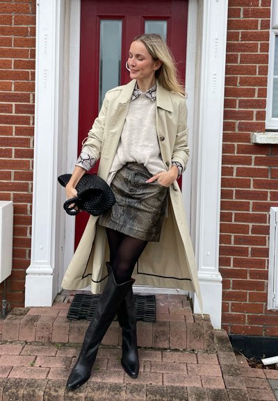 Trench beige sobre un suéter claro, falda con estampado y botas negras hasta la rodilla. Sosteniendo un bolso negro de mimbre, de pie en unos escalones de ladrillo.