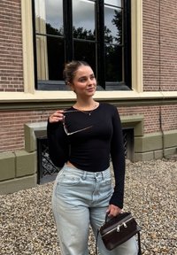 Schwarzes Langarmoberteil, hellblaue hochgeschnittene Jeans, hält eine braune Handtasche und Sonnenbrille. Der Hintergrund zeigt eine Ziegelmauer und Fenster.