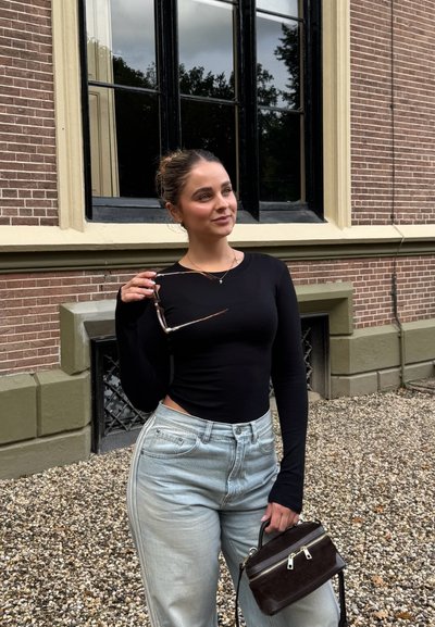 Top negro de manga larga, vaqueros de talle alto en azul claro, sosteniendo un bolso marrón y gafas de sol. El fondo presenta una pared de ladrillos y ventanas.